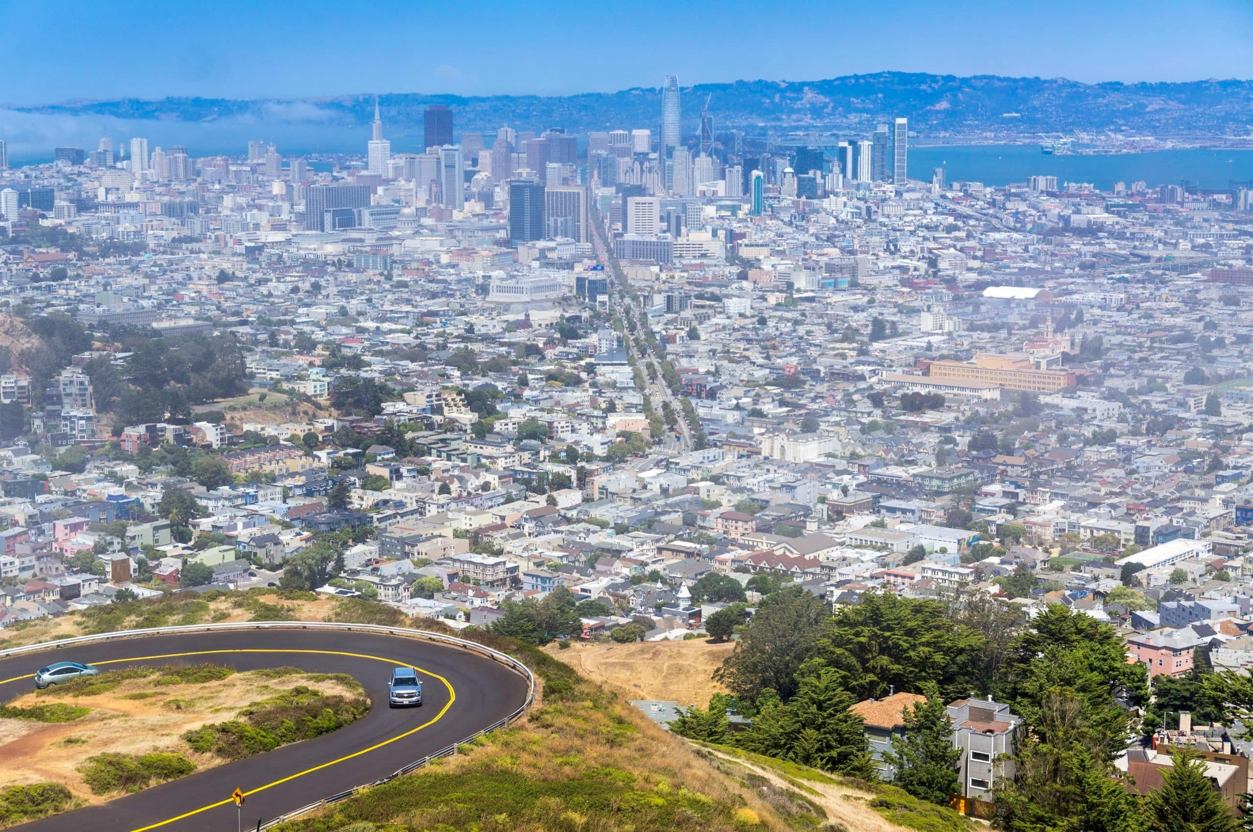La imagen muestra una vista panorámica de la ciudad de San Francisco desde una colina. En primer plano, se observa una carretera en curva que desciende desde lo alto, con un automóvil circulando. En el fondo, se extiende la ciudad, con numerosos edificios, zonas residenciales y una estructura prominente al centro de la imagen.